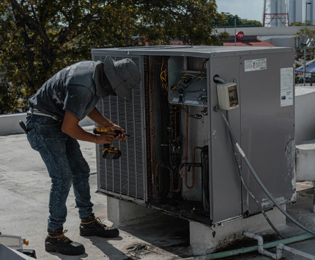 A worker in a bucket hat repairs an outdoor air conditioning unit on a rooftop.